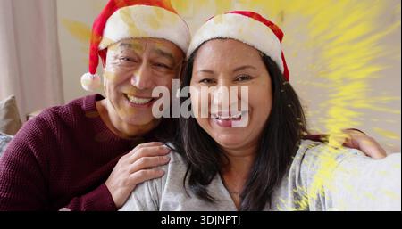 Couple souriant posant sur le canapé, portant des chapeaux de Père Noël, pull marron, haut gris clair, étincelle jaune Banque D'Images