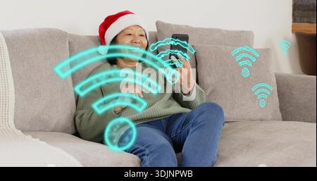 Femme âgée souriante portant chapeau de Père Noël et pull, vérifiant smartphone à la maison avec des icônes Wi ?Fi Banque D'Images
