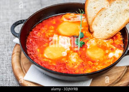 Shakshuka (œufs pochés dans une sauce tomate épicée) avec des toasts croustillants. Il est servi dans une poêle en fonte noire sur un plateau en bois. Gros plan Banque D'Images