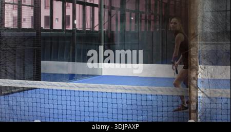 Tenant la pagaie, joueur dans le réservoir noir retournant la balle sur le terrain bleu avec des panneaux de verre, espace de copie. Raquette, femme, padel, tennis, focus, athlétique, sport Banque D'Images