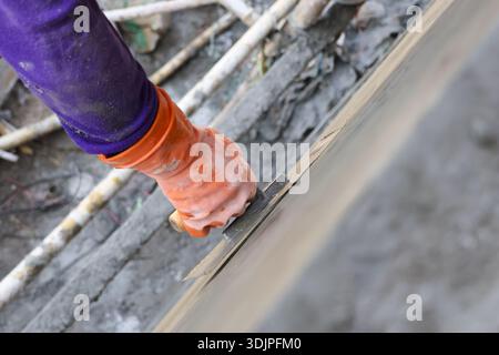 Gros plan de la main de travailleur diligent avec un gant en caoutchouc orange lissant la surface de ciment humide en utilisant l'artisanat précis du chantier de construction occupé à la truelle Banque D'Images