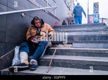 Kiev, Ukraine - 6 mai 2023 : femme âgée avec une jambe bandée et un bâton de marche assis sur les marches de la ville à Kiev, tenant un conteneur, symbolisant le pove Banque D'Images