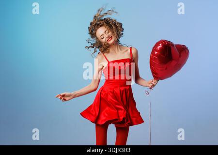Portrait de studio de femme souriante en robe rouge tenant ballon de coeur sur fond bleu avec espace de copie. Convient pour les médias sociaux ou la bannière de site Web dans Banque D'Images