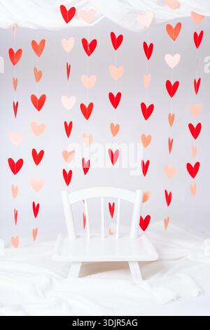 Chaise blanche entourée de décorations de coeur suspendues. Configuration romantique de la Saint-Valentin, scène de photographie d'enfants et fond de célébration d'amour. Banque D'Images