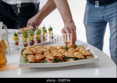 La main d'un homme prend un mini croissant avec remplissage. Gros plan de la nourriture servie lors d'une réception de gala avec croissants et jus dans une cruche Banque D'Images