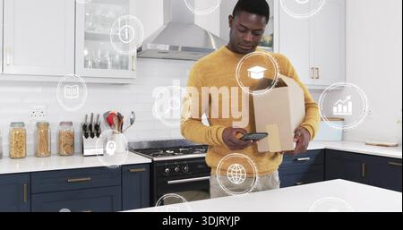 Vérification de l'homme dans le pull moutarde à l'aide d'un smartphone dans la cuisine à la maison, tenant la boîte avec des icônes numériques Banque D'Images