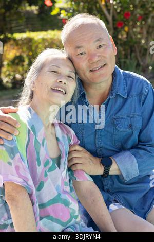 Couple asiatique senior banc assis dans le jardin, penché et souriant, chemise en denim, montre noire visible Banque D'Images