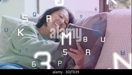 Relaxant femme chinoise senior en pull vert tenant la tablette sur le canapé à la maison avec des lettres flottantes Banque D'Images
