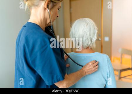 Vue arrière d'une experte médicale examinant le dos d'une femme âgée avec stéthoscope à la clinique Banque D'Images