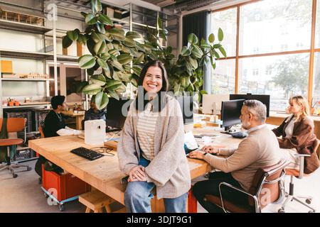 Portrait d'une professionnelle créative féminine souriante assise sur un bureau à proximité de collègues travaillant en arrière-plan au bureau Banque D'Images