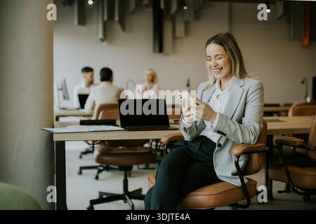Une femme vêtue d'un costume gris clair s'assoit confortablement dans un bureau, en utilisant heureusement son smartphone. En arrière-plan, les collègues travaillent au bureau, créant une prod Banque D'Images