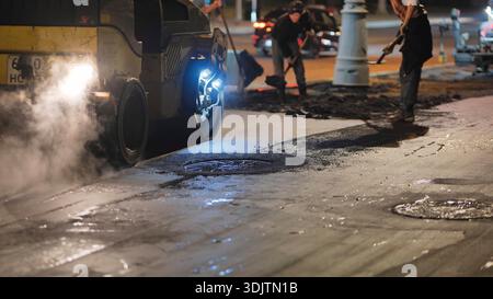 Ouvrier routier exploitant un finisseur d'asphalte la nuit reconstruisant la rue de la ville Banque D'Images
