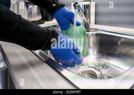Les mains dans des gants bleus rincent une éponge verte sous l'eau courante dans un évier de cuisine élégant, soulignant la propreté et l'importance de l'hygiène dans Banque D'Images