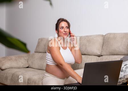 Une femme enceinte sourit en discutant sur son téléphone, confortablement assise sur un canapé moelleux. Son ordinateur portable est ouvert à proximité, mêlant vie de famille et producti Banque D'Images