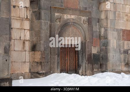 Arménie, Geghard - 10 janvier 2026 : entrée en pierre ornée avec arche sculptée et portes en bois se dresse partiellement entourée de neige au monastère de Geghard Banque D'Images