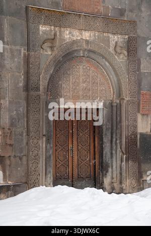 Arménie, Geghard - 10 janvier 2026 : entrée en pierre ornée avec arche sculptée et portes en bois se dresse partiellement entourée de neige au monastère de Geghard Banque D'Images