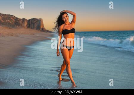 Une femme athlétique asiatique ajustée des îles du Pacifique dans un maillot de bain bikini noir met en valeur son corps tonique tout en souriant sur une plage tropicale. Elle pose chez Go Banque D'Images