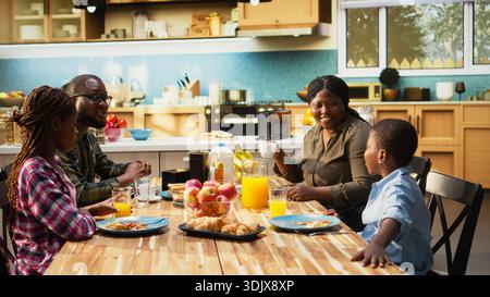 Femme noire versant du jus d'orange dans des verres pour ses enfants et son mari, préparant le repas fait maison et dégustant de la pâtisserie. Les parents et les enfants profitent de leur matinée ensemble, montrant de l'amour. Caméra B. Banque D'Images