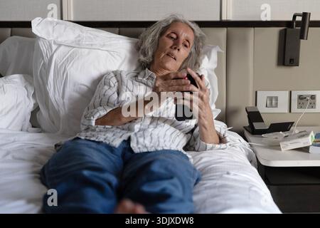 Femme âgée allongée sur un lit en utilisant un smartphone dans une chambre d'hôtel, reflétant les voyages, l'utilisation de la technologie et la vie quotidienne à l'intérieur. Banque D'Images