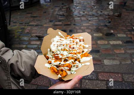 Gros plan de quartiers de pommes de terre chargés avec une sauce blanche crémeuse dans une boîte en carton tenue contre un fond de rue pavé humide dans une ville. Banque D'Images