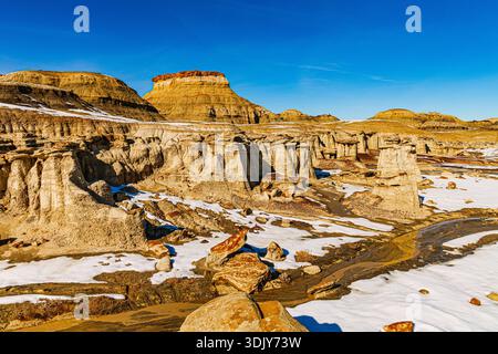 Des formations hoodoo altérées et des falaises de grès érodées entourent des plaques de neige avec une mesa à sommet rouge s'élevant en arrière-plan à Bisti Badlands Banque D'Images