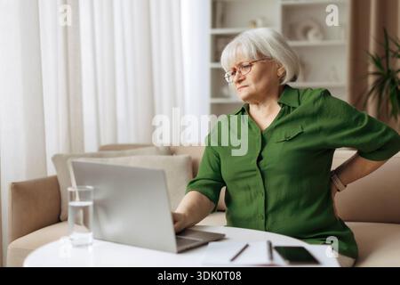 Femme âgée tenant le bas du dos tout en étant assis à l'ordinateur portable à la maison Banque D'Images