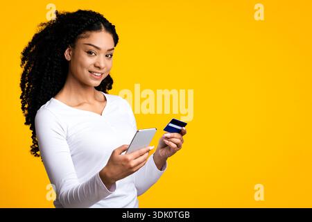 Femme aux cheveux bouclés tient un smartphone et une carte de crédit sur fond jaune Banque D'Images
