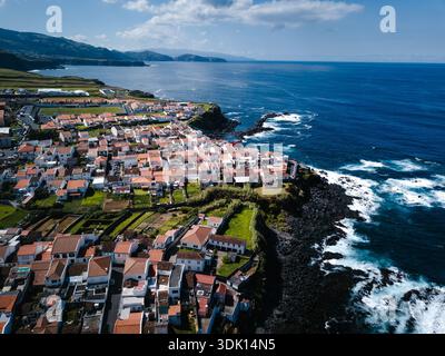Une ville côtière sur les Açores avec des maisons au toit rouge sur des falaises verdoyantes au-dessus de l'océan Atlantique. Les vagues ont frappé la rive rocheuse sous un ciel lumineux, vu d'un Banque D'Images
