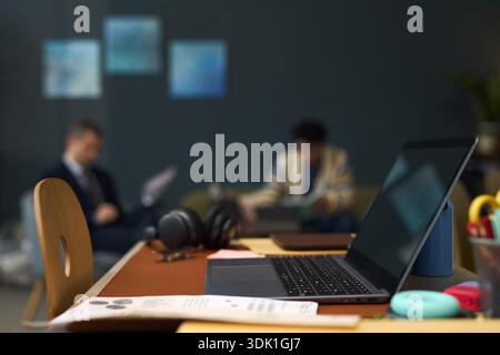 Ordinateur portable ouvert et écouteurs sans fil sur le bureau au premier plan avec deux jeunes hommes adultes flous, un caucasien et un noir, assis en arrière-plan lisant des documents dans un bureau moderne Banque D'Images