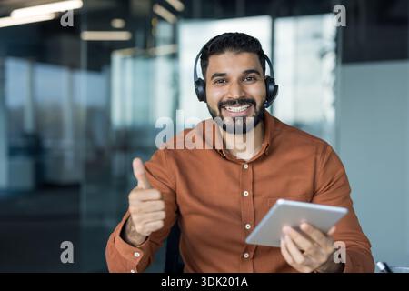 Agent de centre d'appels masculin souriant portant un micro-casque, tenant une tablette numérique et faisant un geste du pouce levé, signifiant un excellent service client et Banque D'Images
