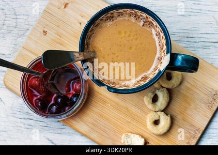 Vue de dessus d'une tasse à café sur une planche de bois avec des biscuits, du beurre, et un pot ouvert de confiture de fruits sauvages avec une cuillère, sur une table légère avec espace de copie. Banque D'Images
