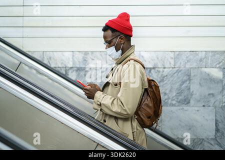Homme voyageur noir se tient sur l'escalator dans le métro ou la gare, porter un masque médical facial pour vous protéger du contact avec le virus de la grippe. Maladies saisonnières, prévention || modèle libéré Banque D'Images