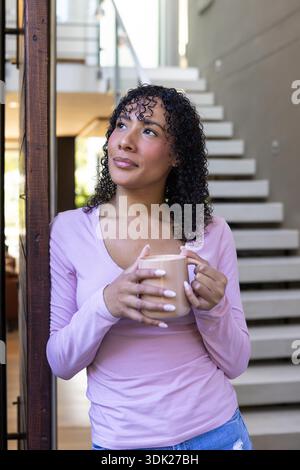 Femme afro-américaine appuyée sur la poutre en bois dans la maison tenant la tasse par escalier blanc regardant vers le haut. Moderne, cosy, minimaliste, détente, architecture, style de vie, intérieur || autorisation du modèle Banque D'Images