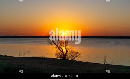 Vue sur le lac tranquille reflète le coucher de soleil ardent illuminant un arbre solitaire à l'horizon, projetant une lueur chaude à travers les eaux calmes, Dallas, Banque D'Images