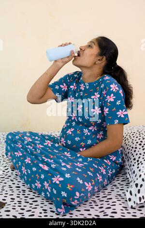 Une femme enceinte est assise sur un lit et boit du lait dans une bouteille dans un cadre intérieur simple. Banque D'Images