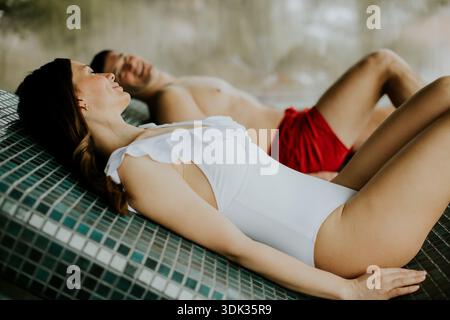 Une femme et un homme se prélassent au bord de la piscine. La femme porte un maillot de bain blanc et sourit en étant allongée sur une surface carrelée. L'homme est en short rouge et apparaît Banque D'Images