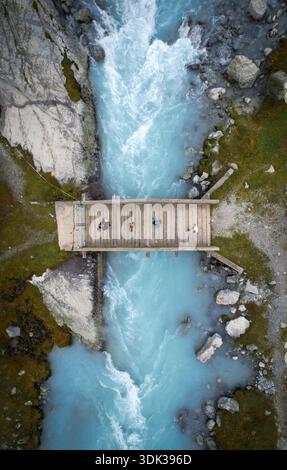 Vue aérienne d'un pont en bois où les gens se tiennent debout au-dessus des eaux bleues laiteuses d'une rivière glaciaire coupant à travers un terrain rocheux, Evolène, Valais, Banque D'Images