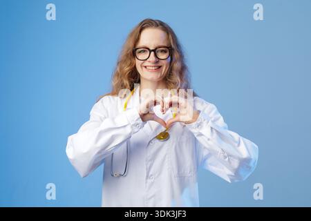 Médecin féminin en manteau blanc avec stéthoscope forme une forme de coeur avec les mains sur fond bleu de studio.concept médical amical pour les lauréats de soins de santé Banque D'Images