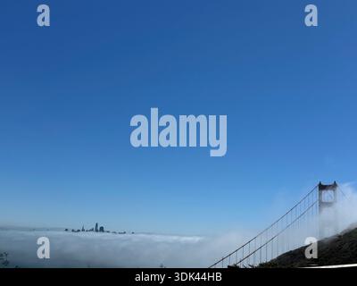 Tour du Golden Gate Bridge s'élevant au-dessus du brouillard côtier avec l'horizon de San Francisco faiblement visible au-delà de la couche marine. Banque D'Images