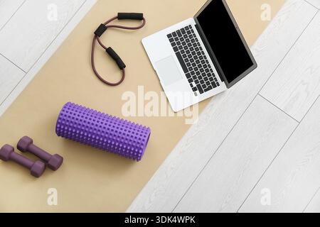 Rouleau en mousse avec ordinateur portable, haltères et extenseur de fitness sur tapis de yoga dans le salon, vue de dessus Banque D'Images