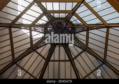 Une vue vers le haut capture un plafond de verre complexe avec des poutres en bois rayonnantes et un moyeu central décoratif en fer forgé, permettant une lumière douce. Banque D'Images
