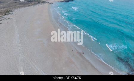 Vue aérienne d'une large plage de sable avec des vagues turquoise de l'océan et de petits groupes de personnes marchant le long du rivage, paysage minimal et calme coasta Banque D'Images