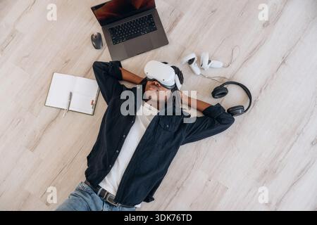 Ordinateur portable à proximité. Vue de dessus de l'homme qui est couché sur le sol dans le casque de réalité virtuelle. Banque D'Images