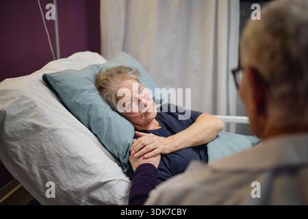 Femme âgée couchée dans le lit d'hôpital tenant les mains avec l'homme d'âge moyen Banque D'Images