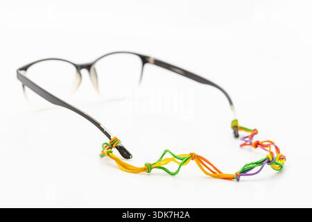 Lunettes noires élégantes avec un bracelet de bricolage créatif fabriqué à partir de bandes de caoutchouc colorées liées sur un fond blanc. Accessoire de lunettes fait main unique Banque D'Images