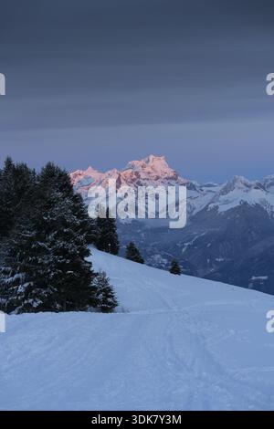 Vue du premier plan enneigé menant aux montagnes lointaines, ensoleillées et enneigées sous un ciel sombre, Morgins, Valais, Suisse. Banque D'Images