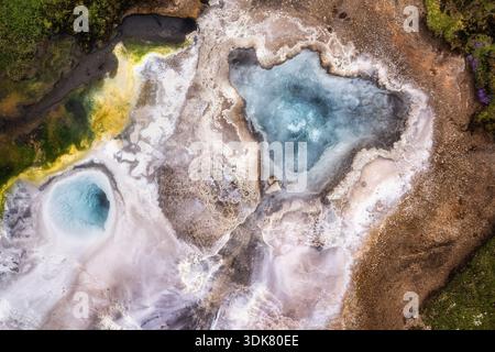 La vue aérienne des piscines géothermiques scintille de bleus éthérés au milieu d'un paysage de bruns terreux et de verts verdoyants, créant une tapisserie surréaliste de nat Banque D'Images