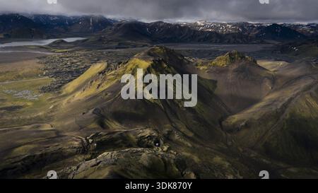 Vue aérienne des montagnes escarpées s'élèvent avec des taches de mousse verte vibrante contre le terrain sombre et rocheux, contrastant avec les sommets enneigés lointains, S. Banque D'Images