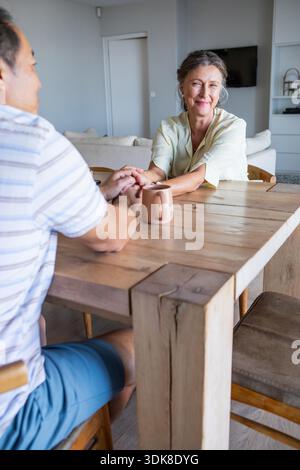 Couple diversifié tenant les mains sur la table à manger en bois dans le salon avec tasse en céramique. Romantique, intimité, connexion, confortable, moderne, style de vie, chaleur || autorisation du modèle Banque D'Images