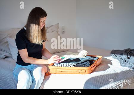 Femme assise sur le lit mettant des objets personnels dans la valise ouverte dans la chambre. Préparation et emballage du voyage Banque D'Images